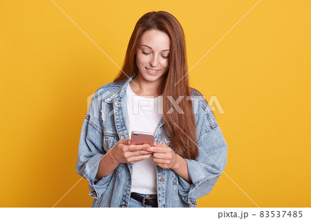 Portrait of happy beautiful young woman with dark straight hair texting message on her phone and smiling, checking social network, wearing denim jacket, posing isolated over yellow background. Portrait of happy beautiful young woman with dark straight hair texting message on her phone and smiling, checking social network, wearing denim jacket, posing isolated over yellow background. 83537485
