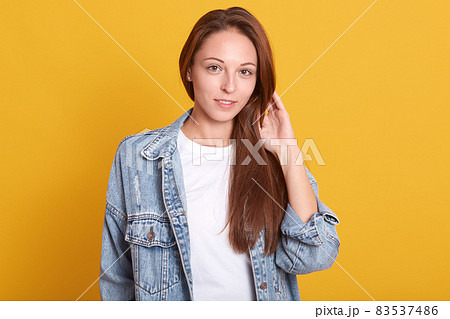 Horizontal shot of young charming woman in denim jacket and white casual shirt standing over yellow background, touches her beautiful long hair, looking directly at camera, being in good mood. 83537486