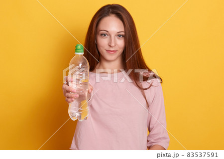 Portrait of beautiful sports woman showing bottle of water isolated on yellow background, girl wearing pink shirt, attractive famale having long brown hair, lady looking smiling directly at camera. Portrait of beautiful sports woman showing bottle of water isolated on yellow background, girl wearing pink shirt, attractive famale having long brown hair, lady looking smiling directly at camera. 83537591
