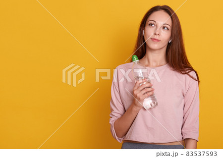 Young athletic woman wearing wireless headphones and rose sportswear holds bottle of water in hands, looking up, ready for running, isolated over yellow wall, copy space for promotion or advertismant. Young athletic woman wearing wireless headphones and rose sportswear holds bottle of water in hands, looking up, ready for running, isolated over yellow wall, copy space for promotion or advertismant. 83537593