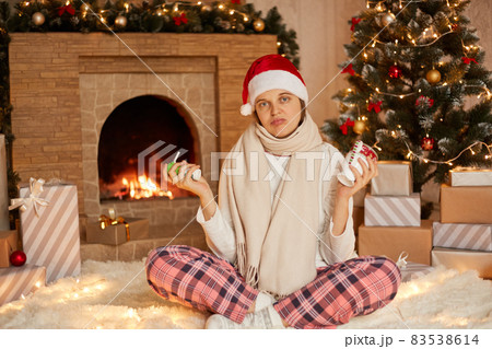 Young sick girl in santa hat and checkered pants, holding spray and cup of warming drink in hands, looks at camera with sad facial expression, being in during Christmas holidays. 83538614