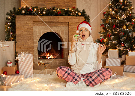 Woman having problem with health, being at home, lady sitting in floor with crossed legs, showing thermometer to camera, using sore throat spray, wearing home closing and santa hat. 83538626