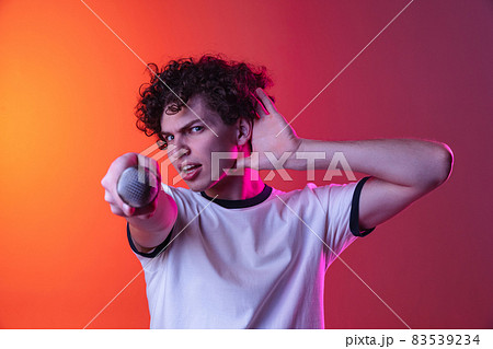 Cropped portrait of young man in white T-shirt directing microphone on camera isolated over gradient pink orange background in neon lights Cropped portrait of young man in white T-shirt directing microphone on camera isolated over gradient pink orange background in neon lights 83539234