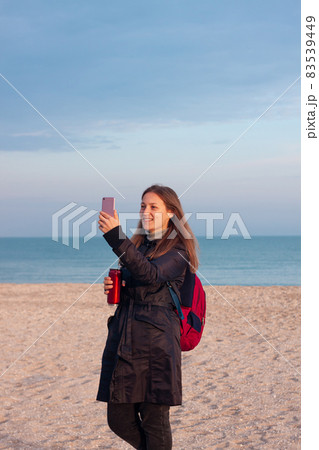 Happy young woman in black raincoat empty autumn sea beach. Smiling girl drinking tea with thermo can bottle. Lifestyle Happy young woman in black raincoat empty autumn sea beach. Smiling girl drinking tea with thermo can bottle. Lifestyle 83539449
