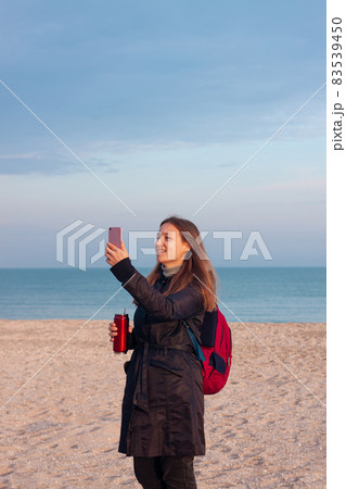Happy young woman in black raincoat empty autumn sea beach. Smiling girl drinking tea with thermo can bottle. Lifestyle Happy young woman in black raincoat empty autumn sea beach. Smiling girl drinking tea with thermo can bottle. Lifestyle 83539450