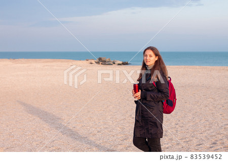 Happy young woman in black raincoat empty autumn sea beach. Smiling girl drinking tea with thermo can bottle. Lifestyle 83539452