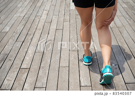 Rear view of unrecognizble female runner in blue sports shoes standing on wooden pier and touching pain point while feeling pain under kneecap 83540307