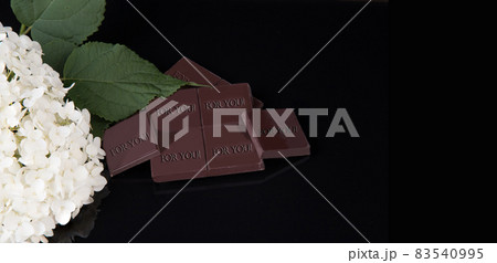 A broken chocolate bar with the inscription FOR YOU lies on a black mirror background and a branch of a white hydrangea. Copy Space 83540995