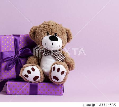 cute brown teddy bear and box wrapped in paper and silk ribbon on a purple background. Prize and congratulations cute brown teddy bear and box wrapped in paper and silk ribbon on a purple background. Prize and congratulations 83544535