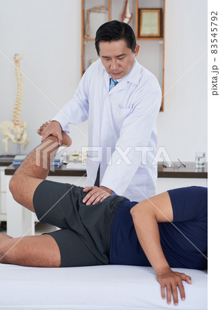 Vertical medium long shot of mature Asian physical therapist wearing white coat working with unrecognizable male patient recovering his hip joints range of motion Vertical medium long shot of mature Asian physical therapist wearing white coat working with unrecognizable male patient recovering his hip joints range of motion 83545792