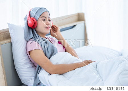 Happy a asian women disease mammary cancer patient with pink ribbon wearing headscarf in headphones is listening to music After treatment to chemotherapy sit on bed In the bedroom at the house 83546151