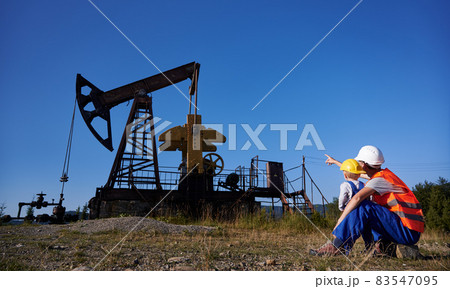 Back view of engineer pointing index finger at petroleum borehole pump in the oil field to preschool boy. Adult, acting mechanic and his little future successor observing the operation of oil pump. Back view of engineer pointing index finger at petroleum borehole pump in the oil field to preschool boy. Adult, acting mechanic and his little future successor observing the operation of oil pump. 83547095