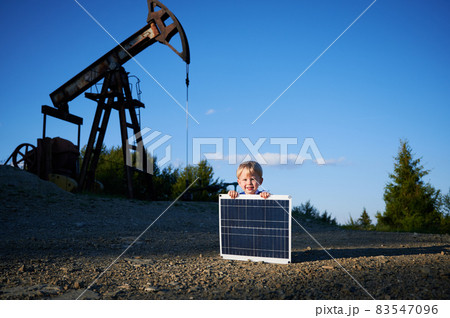 Portrait of cheerful boy with solar battery smiling at camera during education time at petroleum station with pump derrick on background, happy child with panel for product alternative green energy 83547096