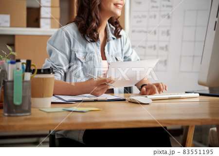 Cropped image of smiling secretary sitting at table and holding document while entering data into computer 83551195