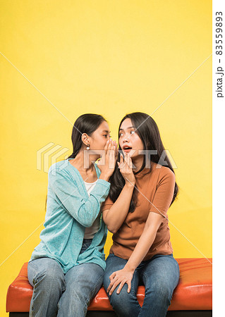 a beautiful woman is surprised when her friend whispers while chatting sitting together on the couch a beautiful woman is surprised when her friend whispers while chatting sitting together on the couch 83559893
