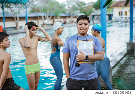 a swimming trainer smiling with one hand in pocket while holding a clipboard with his students 83559938