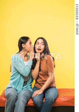 a young asian woman with a surprised gesture as her friend whispers while sitting together a young asian woman with a surprised gesture as her friend whispers while sitting together 83560013