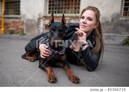 A girl in leather, with a Doberman, in an old yard. 83560720