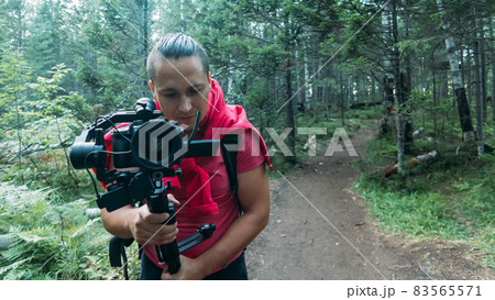 Professional Videographer Holding DSLR Camera on 3-axis Gimbal Stabilization Device in Forest. Cinematographer Operator. Slow Motion Professional Videographer Holding DSLR Camera on 3-axis Gimbal Stabilization Device in Forest. Cinematographer Operator. Slow Motion 83565571