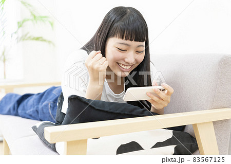 自宅のソファで寝そべってスマホでネットテレビを観て熱狂する若い女性 自宅のソファで寝そべってスマホでネットテレビを観て熱狂する若い女性 83567125
