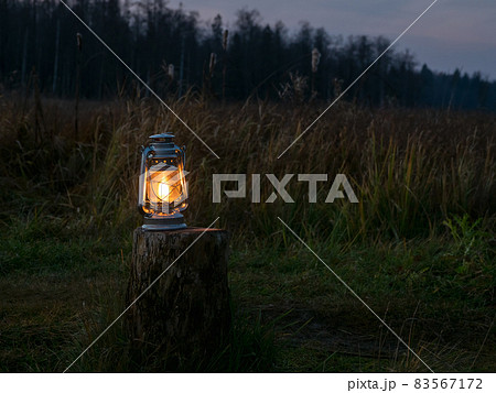 An old oil lantern with a lit fire stands on the stump outdoors at night. Soft warm yellow light of an old kerosene lamp. 83567172