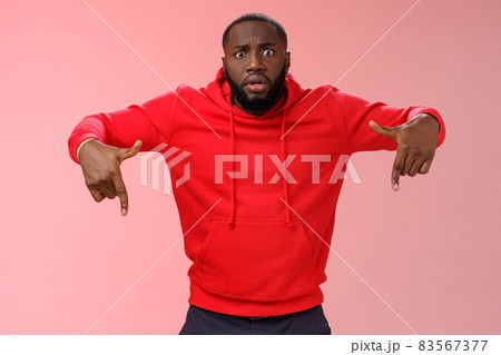 Nervous upset gasping young bearded african american man widen eyes shocked pointing down lose hope, standing worried anxious losing pari, wearing red hoodie pink background 83567377
