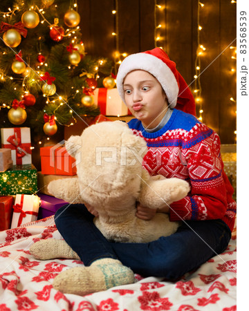 Child girl posing in new year or christmas decoration. Festive lights and lots of gifts, an elegant Christmas tree with toys. The girl is wearing a red sweater and a Santa hat, she plaing with bear 83568539