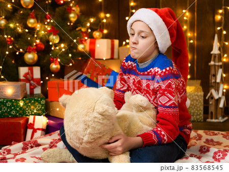 Child girl posing in new year or christmas decoration. Festive lights and lots of gifts, an elegant Christmas tree with toys. The girl is wearing a red sweater and a Santa hat, she plaing with bear 83568545