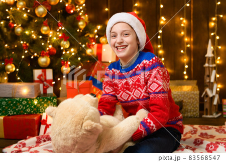 Child girl posing in new year or christmas decoration. Festive lights and lots of gifts, an elegant Christmas tree with toys. The girl is wearing a red sweater and a Santa hat, she plaing with bear 83568547