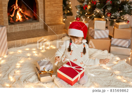 Cute little girl opening Christmas gift at home while sitting on soft carpet on floor, looking at box, pulls ribbon to open present, dresses white pullover and red santa hat. 83569862