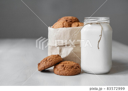 Milk in a glass jar and oatmeal cookies in a paper bag on a white table background. Copy, empty space for text Milk in a glass jar and oatmeal cookies in a paper bag on a white table background. Copy, empty space for text 83572999