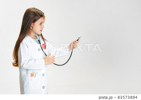Close-up portrait of cute little girl, child in image of doctor wearing white lab coat with stethoscope isolated on white studio background Close-up portrait of cute little girl, child in image of doctor wearing white lab coat with stethoscope isolated on white studio background 83573894