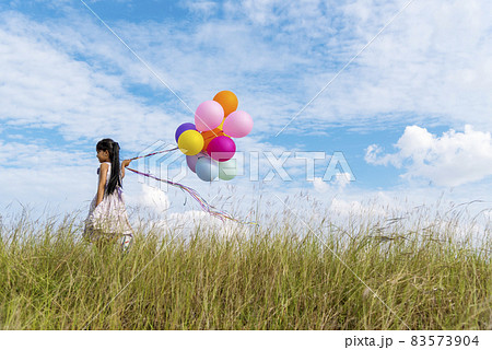 Cheerful cute girl holding balloons running on green meadow white cloud and blue sky with happiness. Hands holding vibrant air balloons play on birthday party happy times summer on sunlight outdoor 83573904