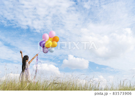 Cheerful cute girl holding balloons running on green meadow white cloud and blue sky with happiness. Hands holding vibrant air balloons play on birthday party happy times summer on sunlight outdoor 83573906