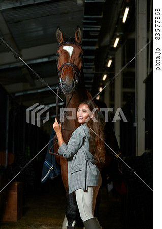 Portrait of smiling female jockey standing by horse in stable 83577363