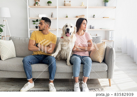 Unhappy offended young diverse couple looking in opposite directions, sitting on sofa with cute dog between them Unhappy offended young diverse couple looking in opposite directions, sitting on sofa with cute dog between them 83582939
