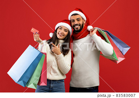 Christmas sales and shopping. Arab couple with shopper bags and credit card, posing in Santa hats on red background 83583007