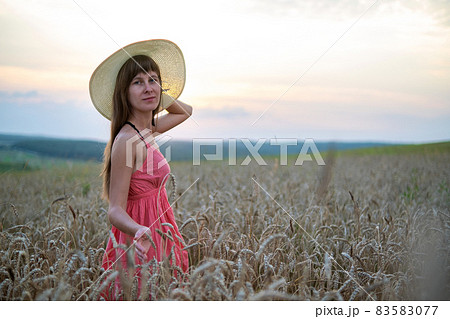 Young happy woman in red summer dress and white straw hat walking on yellow farm field with ripe golden wheat enjoying warm evening. 83583077