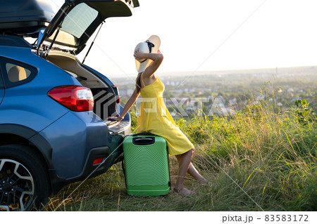 Young woman resting on a green suitcase near her car in summer nature. Travel and vacations concept. Young woman resting on a green suitcase near her car in summer nature. Travel and vacations concept. 83583172