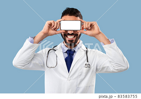 Male Doctor Holding Smartphone In Front Of Eyes, Blue Background Male Doctor Holding Smartphone In Front Of Eyes, Blue Background 83583755