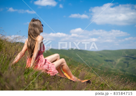 Young woman in red dress resting on green grassy field on a sunny day in summer mountains enjoying view of nature. 83584715