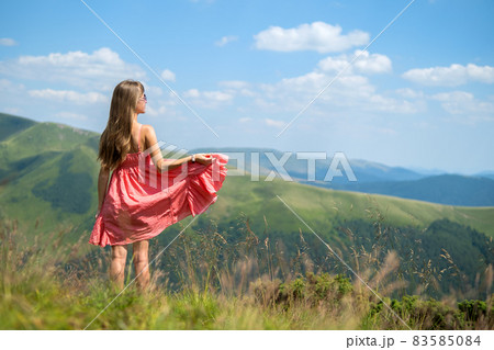Young woman in red dress standing on grassy field on a windy day in summer mountains enjoying view of nature. 83585084