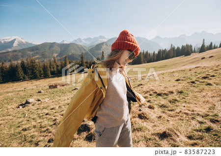 Young blond woman with sunglasses dancing in mountains. Freedom, happiness, travel and vacations concept, outdoor activities. Happy Girl wearing red hat and a yellow jacket. Wind in the hair. 83587223