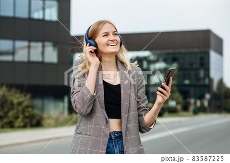 Portrait of young happy blonde woman listening to music with headphones and smiling while walking on the street in the city. Music lover enjoying music. Portrait of businesswoman walking Portrait of young happy blonde woman listening to music with headphones and smiling while walking on the street in the city. Music lover enjoying music. Portrait of businesswoman walking 83587225