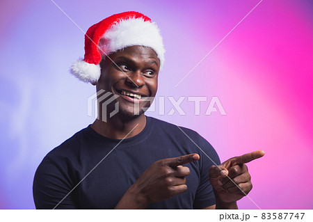 Portrait of positive african american smiling man in santa hat and casual t-shirt on colourful background, christmas time. Winter holidays and emotional people concept. 83587747