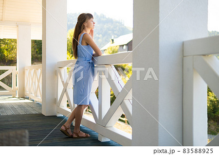 Side view of young woman in blue dress looking on nature on balcony. Concept of enjoying fresh air and good weather. 83588925