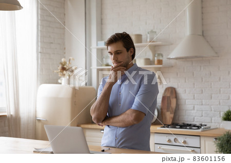 Man standing in kitchen staring at laptop thinks over task 83601156