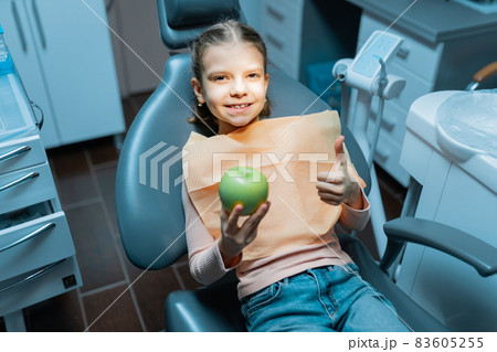 Child girl sits in dental chair gestures thumb up while holds apple. treatment, medicine, dentistry 83605255