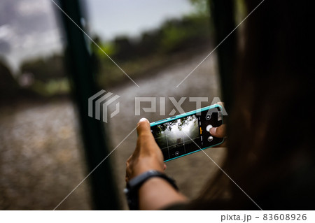 Rear view of a young woman making picture on phone camera while enjoying her vacation holidays in the rainy forest, girl photographing amazing landscape on her smart phone 83608926