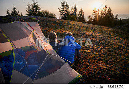 Back view of two travelers man and woman sitting on grass in front of tent set up on mountain hill, early in the morning and contemplating on sunny sky. Concept of hiking, camping and relationships. 83613704